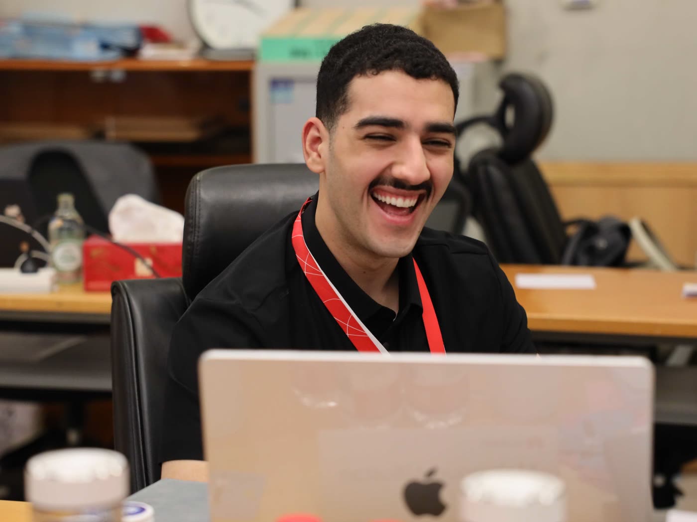 Mahmoud Albustan laughing at a desk, a MacBook open in front of him